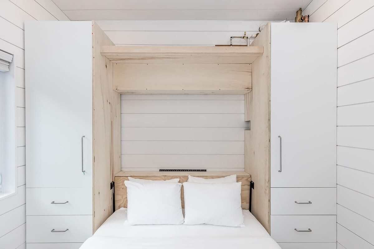 A bedroom with a bed, two white pillows, and a wooden headboard. There are two white wardrobes with silver handles.