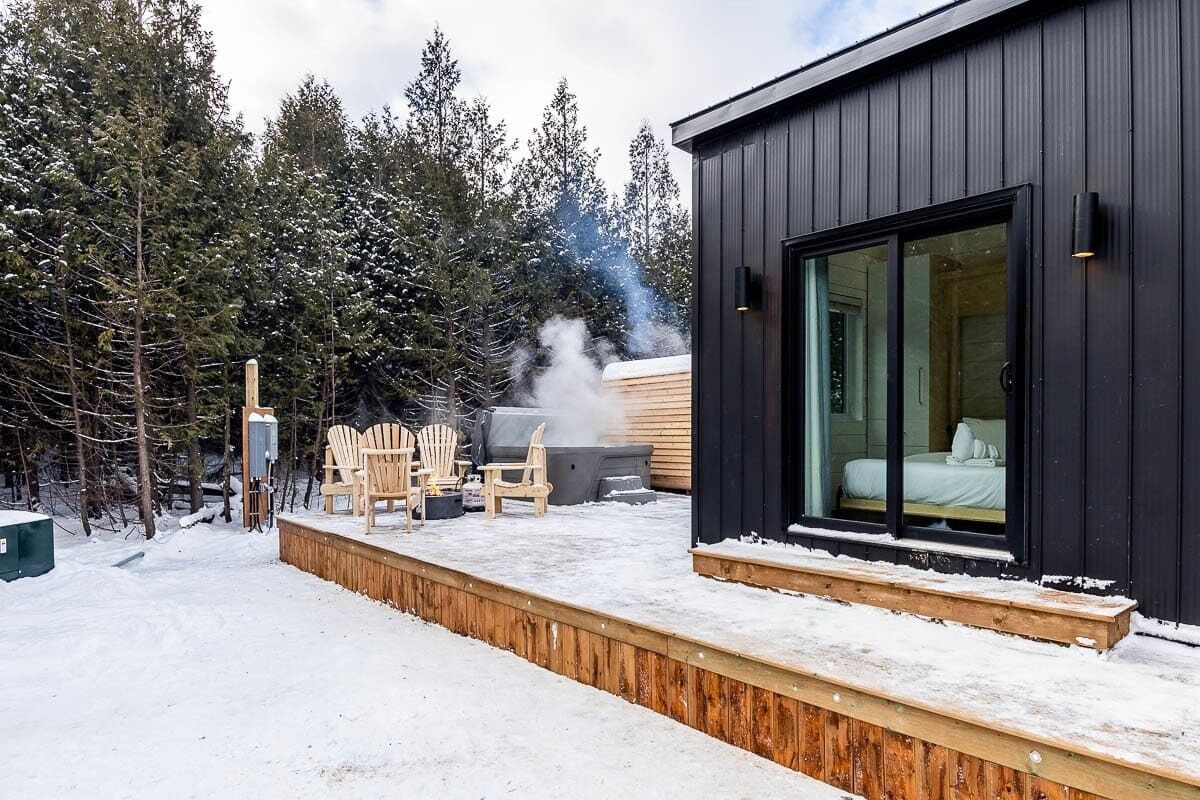 Una cabaña moderna con terraza, mobiliario exterior y un dormitorio a la vista con un ventanal grande.