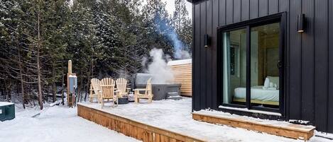 Una cabaña moderna con terraza, mobiliario exterior y un dormitorio a la vista con un ventanal grande.