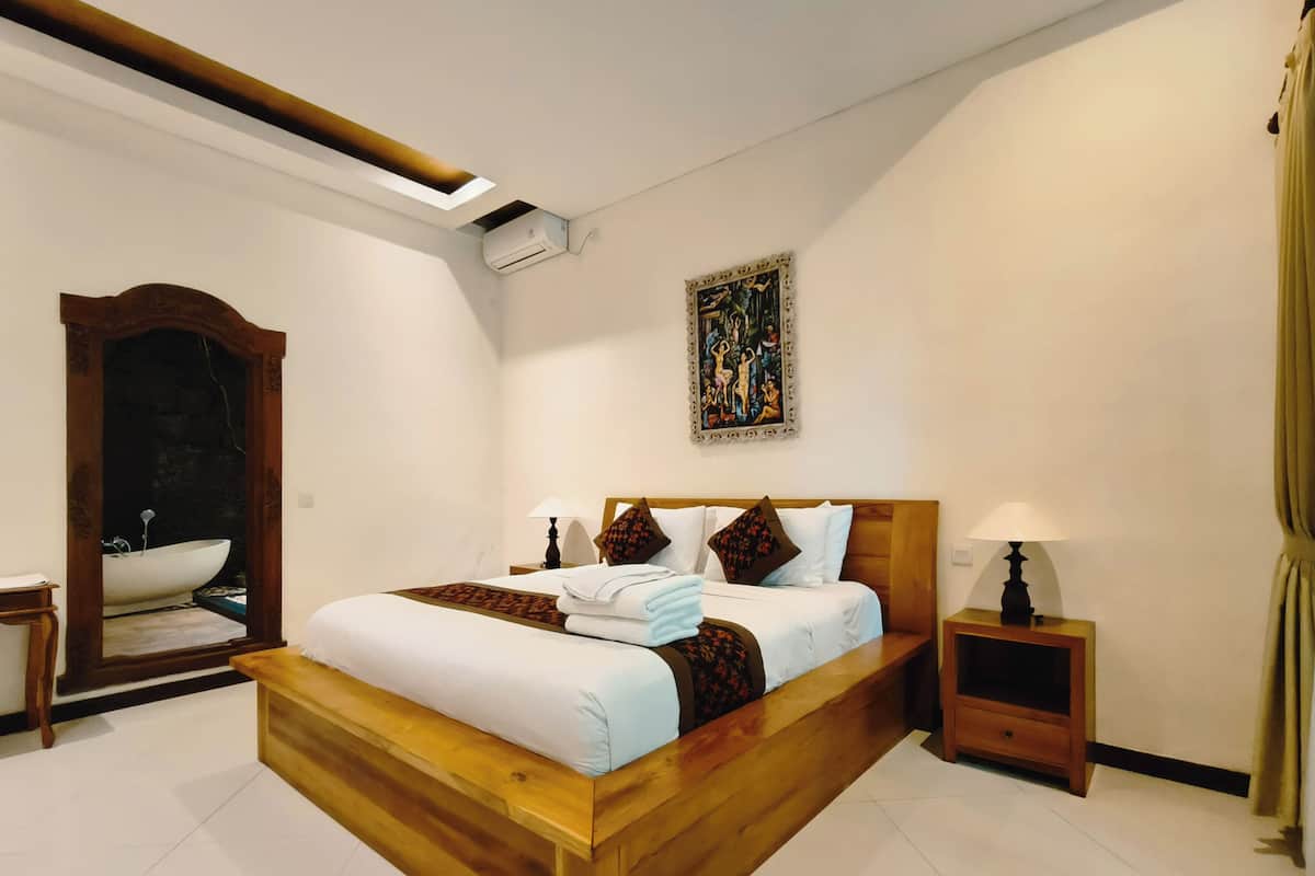 A modern hotel room with a large bed, a wooden headboard, a nightstand with a lamp, a painting on the wall, and a bathroom with a bathtub visible through an archway.