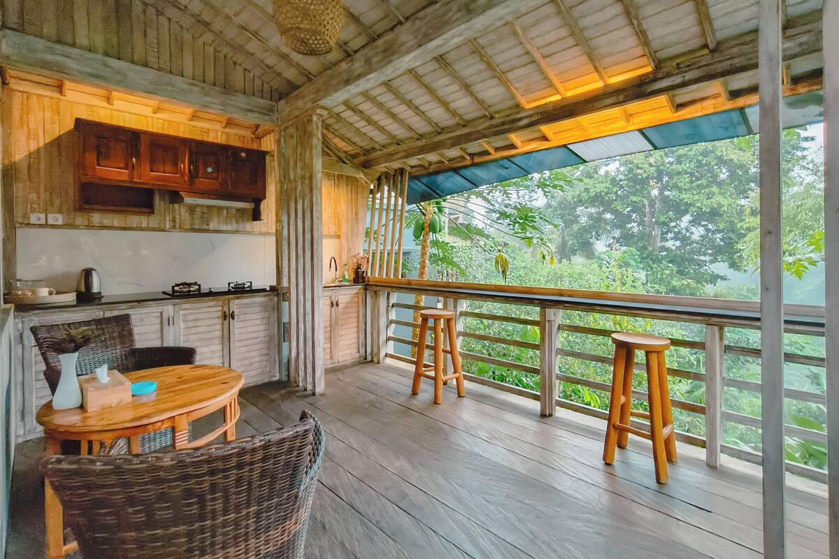 A wooden deck with a table and chairs, a hanging light fixture, and a view of greenery.
