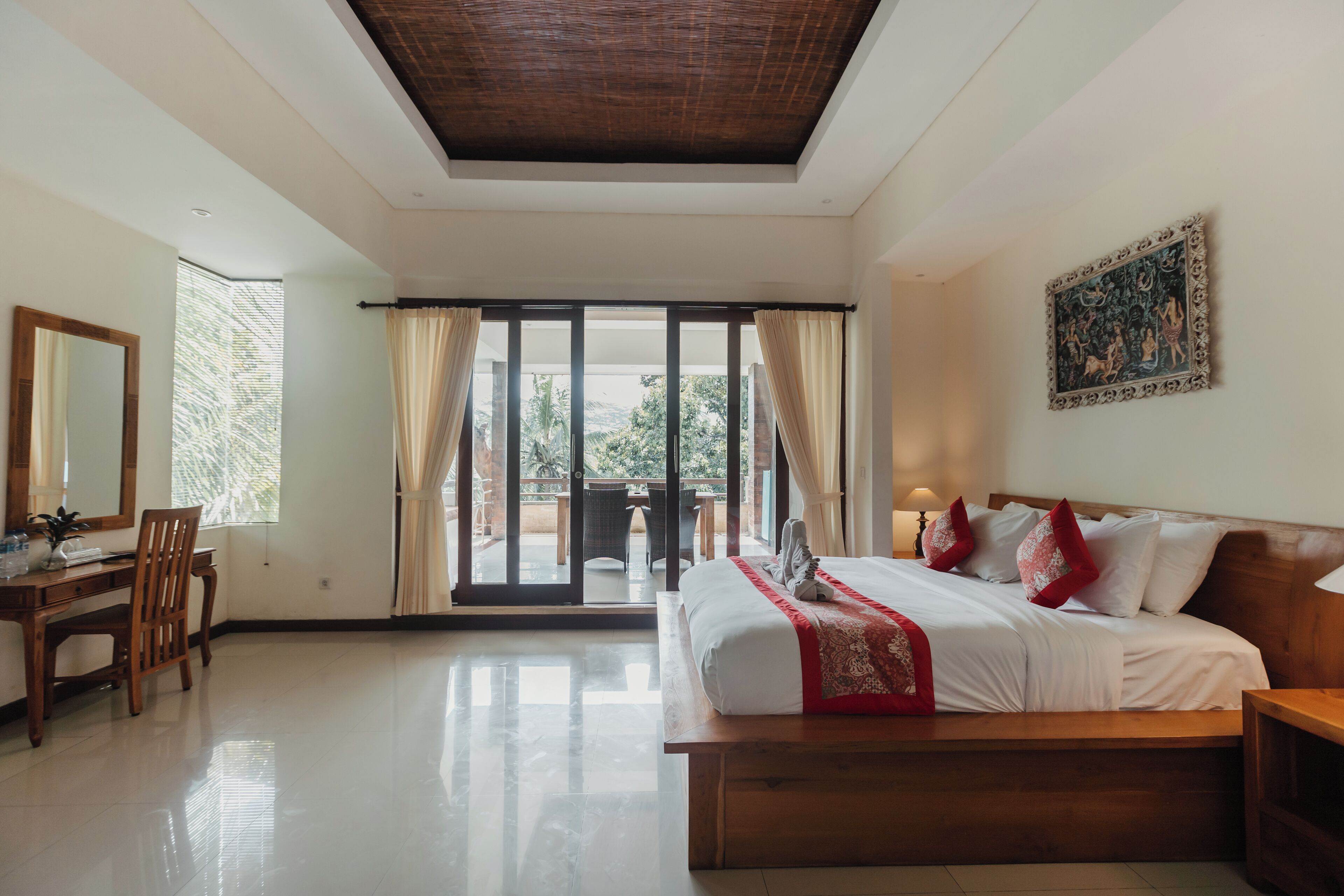 Quarto com uma cama grande, uma escrivaninha de madeira e uma cadeira. Há uma porta de vidro deslizante que dá para uma área externa.