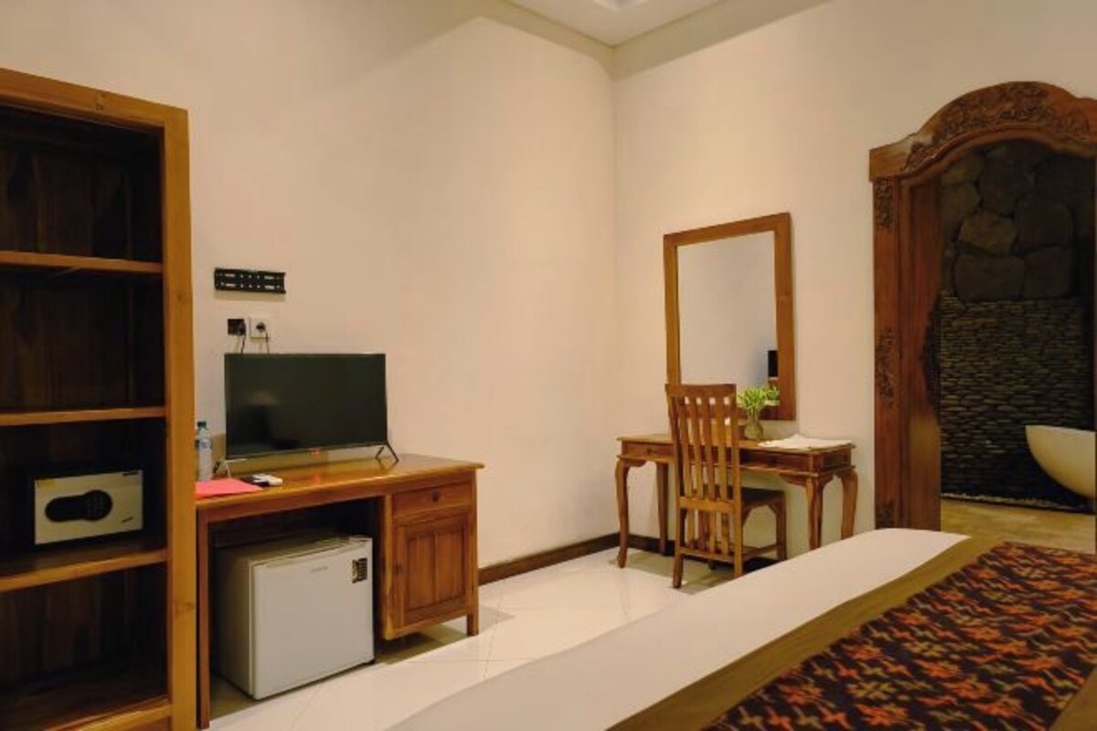 A hotel room with a wooden desk, a television, a small refrigerator, a chair, a mirror, and a stone wall.
