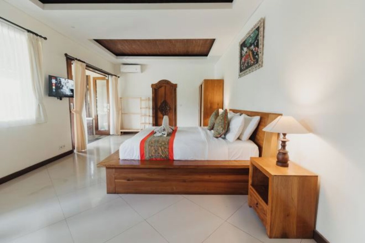 A bedroom with a wooden bed, a nightstand with a lamp, and a television on the wall.