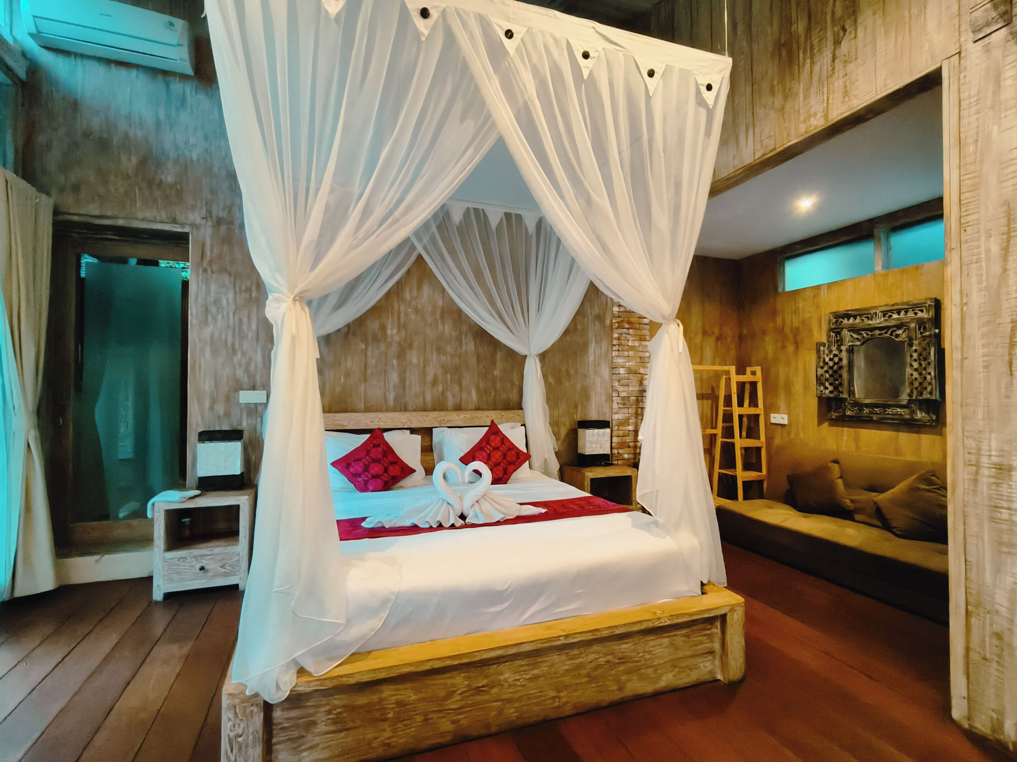 A four-poster canopy bed with white bedding and red pillows, a wooden headboard, and a bedside table with a lamp.