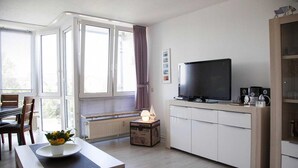 A modern living room with a large window, a flat-screen TV, a dining table with chairs, and a small cabinet. - Ferienwohnung in Cuxhaven-Duhnen (Cuxhaven)