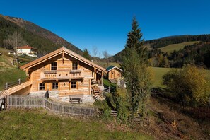 A log cabin with a balcony, surrounded by a wooden fence, set in a mountainous area with a clear sky.