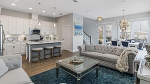 Una cocina moderna con armarios blancos, una isla central con taburetes, un comedor con candelabro y una sala de estar con sofá y mesa de centro.