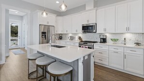 Una cocina moderna con armarios blancos, una isla grande con taburetes de bar, electrodomésticos de acero inoxidable y un salpicadero de baldosas en patrón de espiga.