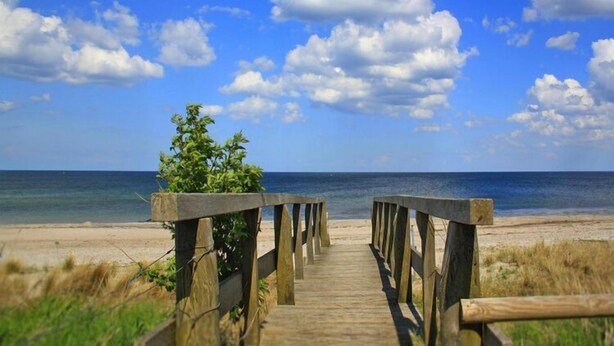 A wooden boardwalk leading to a beach with a clear view of the ocean and a partly cloudy sky.