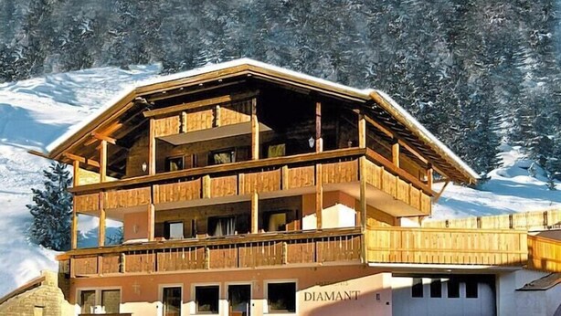 Een houten chalet met meerdere verdiepingen en balkons, genaamd “DIAMANT”, gelegen tegen een besneeuwd berglandschap.