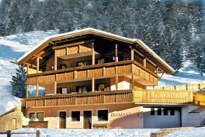 Eine mehrstöckige Holzhütte mit Balkonen, mit dem Schriftzug „DIAMANT“ versehen, vor einer verschneiten Berglandschaft.
