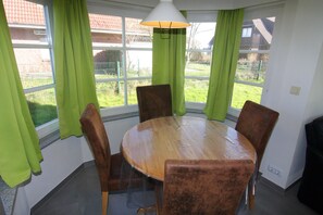 Un coin repas avec une table ronde en bois et quatre chaises, de grandes fenêtres avec des rideaux verts et une lampe suspendue.