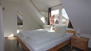 Une chambre avec un plafond en pente, une grande fenêtre, un lit avec une literie blanche, une table de chevet avec une lampe et un miroir sur le mur.