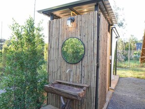 Eine hölzerne Außentoilette mit einem runden Spiegel, einem Waschbecken und einem Metallarmaturen.