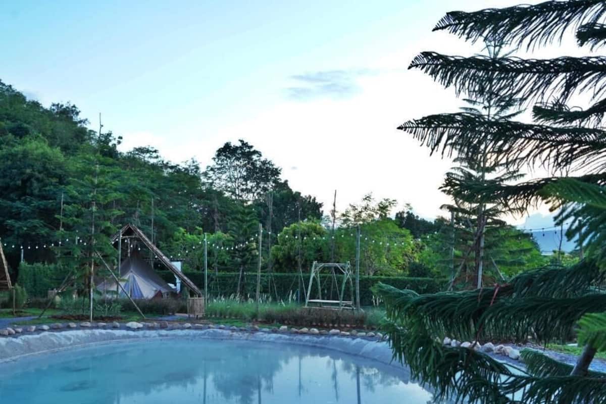 Ein friedlicher Poolbereich im Freien mit freiem Blick auf den Himmel, umgeben von üppigem Grün und einer kleinen Struktur, die wie ein Gazebo oder Spielbereich wirkt.