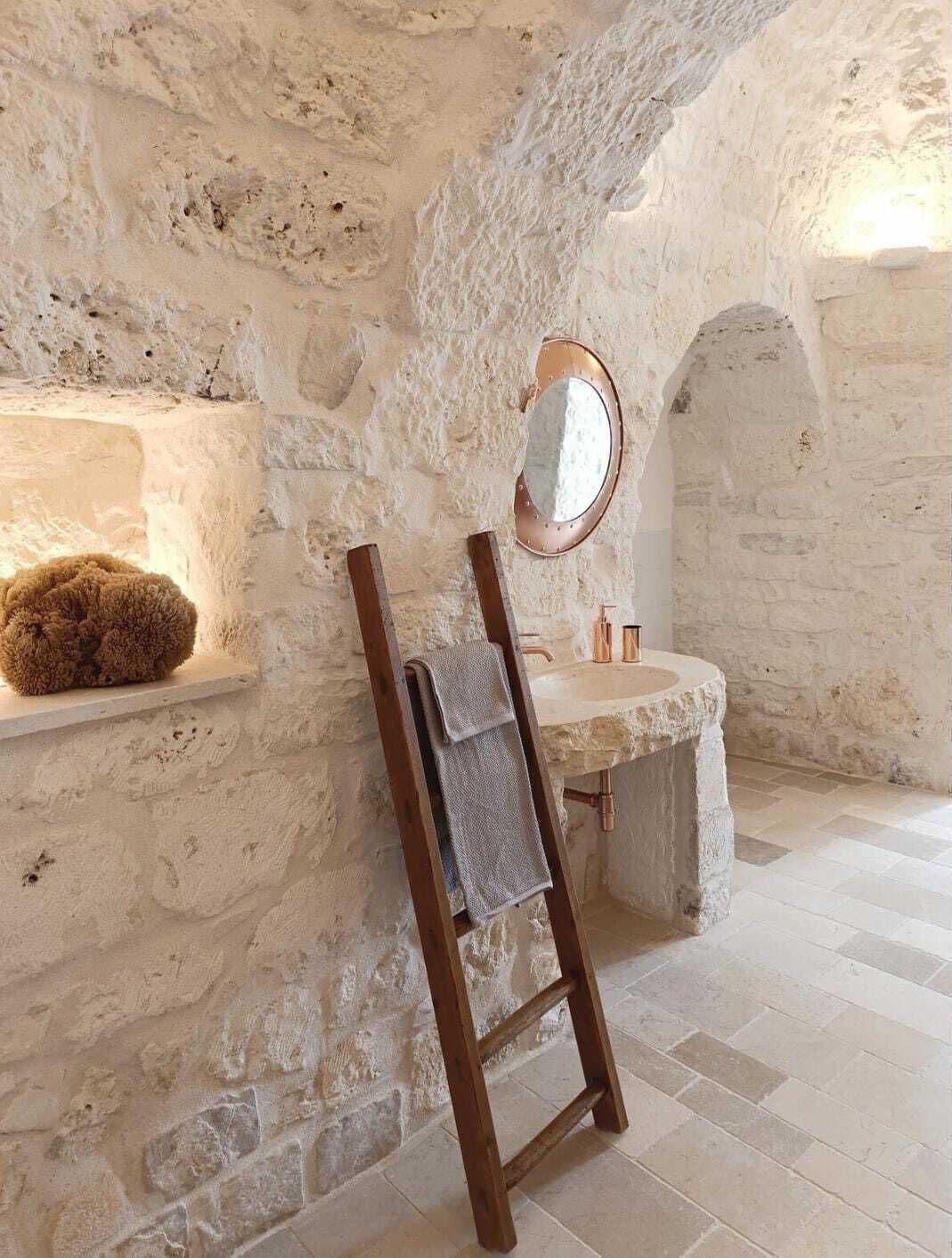 Une salle de bain aux murs en pierre, avec une échelle en bois, un miroir rond et un lavabo beige.