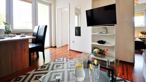Een moderne woonkamer met een houten eettafel, een zwarte lederen fauteuil, een glazen salontafel en een kamerbreed tapijt met patroon. Een flatscreen televisie is aan de muur bevestigd en er staat een witte boekenkast met decoratieve objecten.