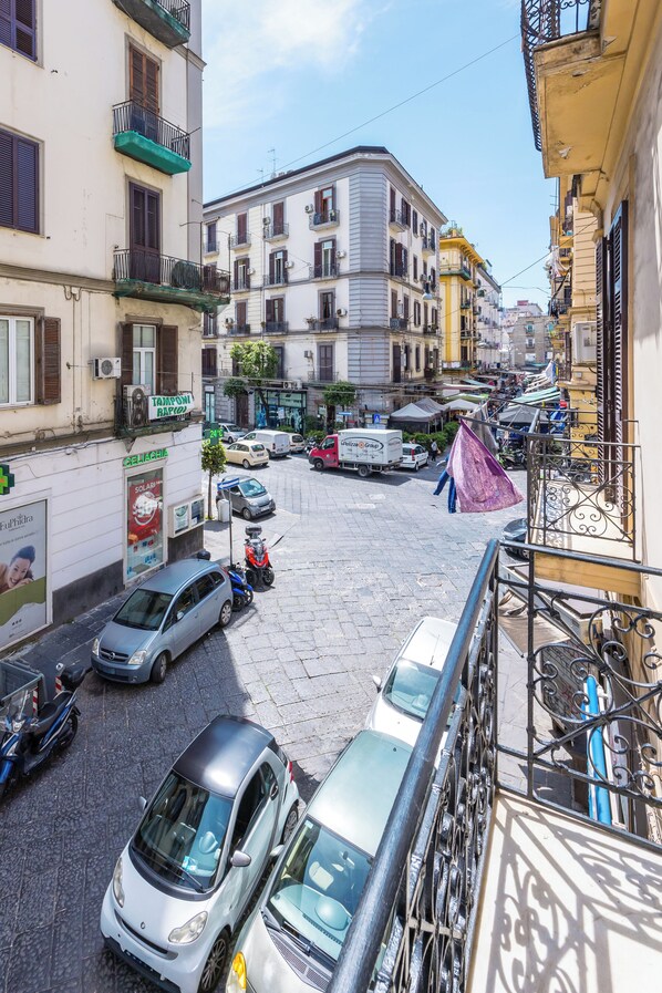 Una calle estrecha con coches aparcados y una moto, edificios con balcones y comercios, bajo un cielo despejado.