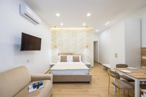 Una habitación de hotel moderna con cama, mesitas de noche, televisor, sofá, mesa de comedor con sillas y aire acondicionado.