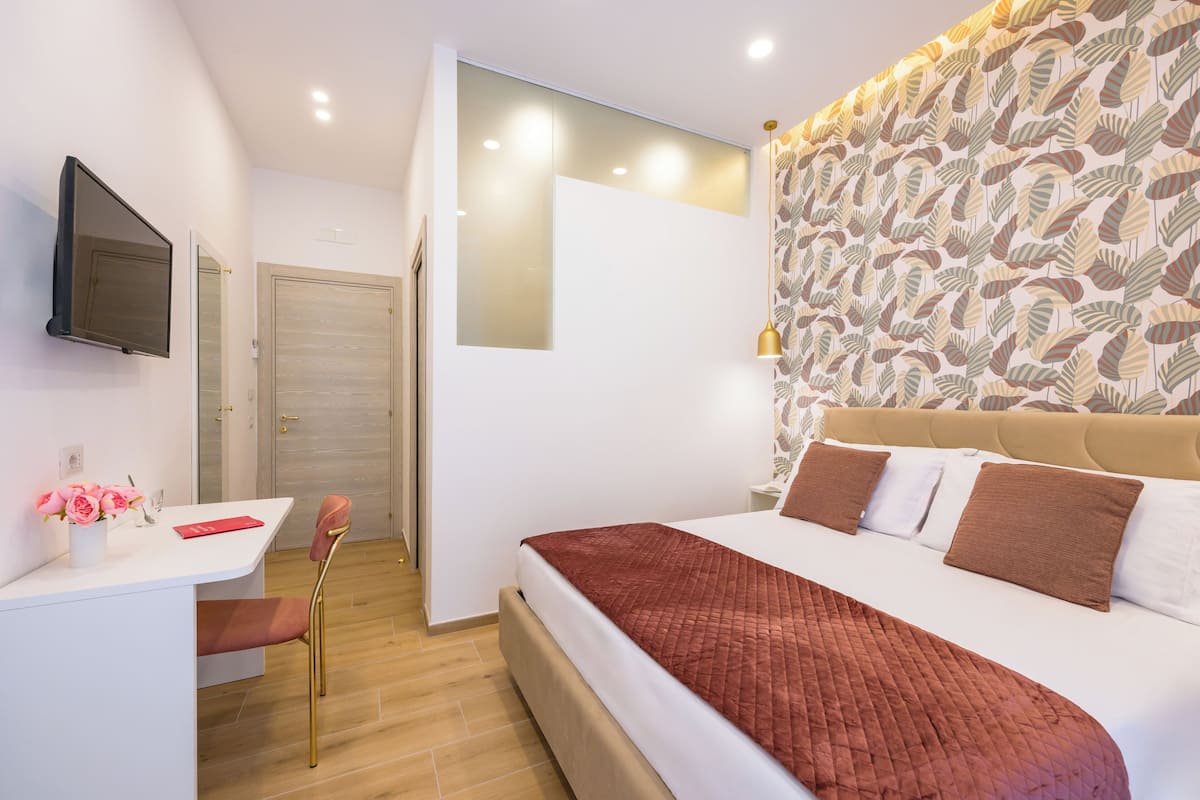 Moderni hotellihuone, jossa on suuri sänky, työpöytä tuolilla, seinälle asennettu televisio ja kuviollinen tapetti seinällä sängyn takana.