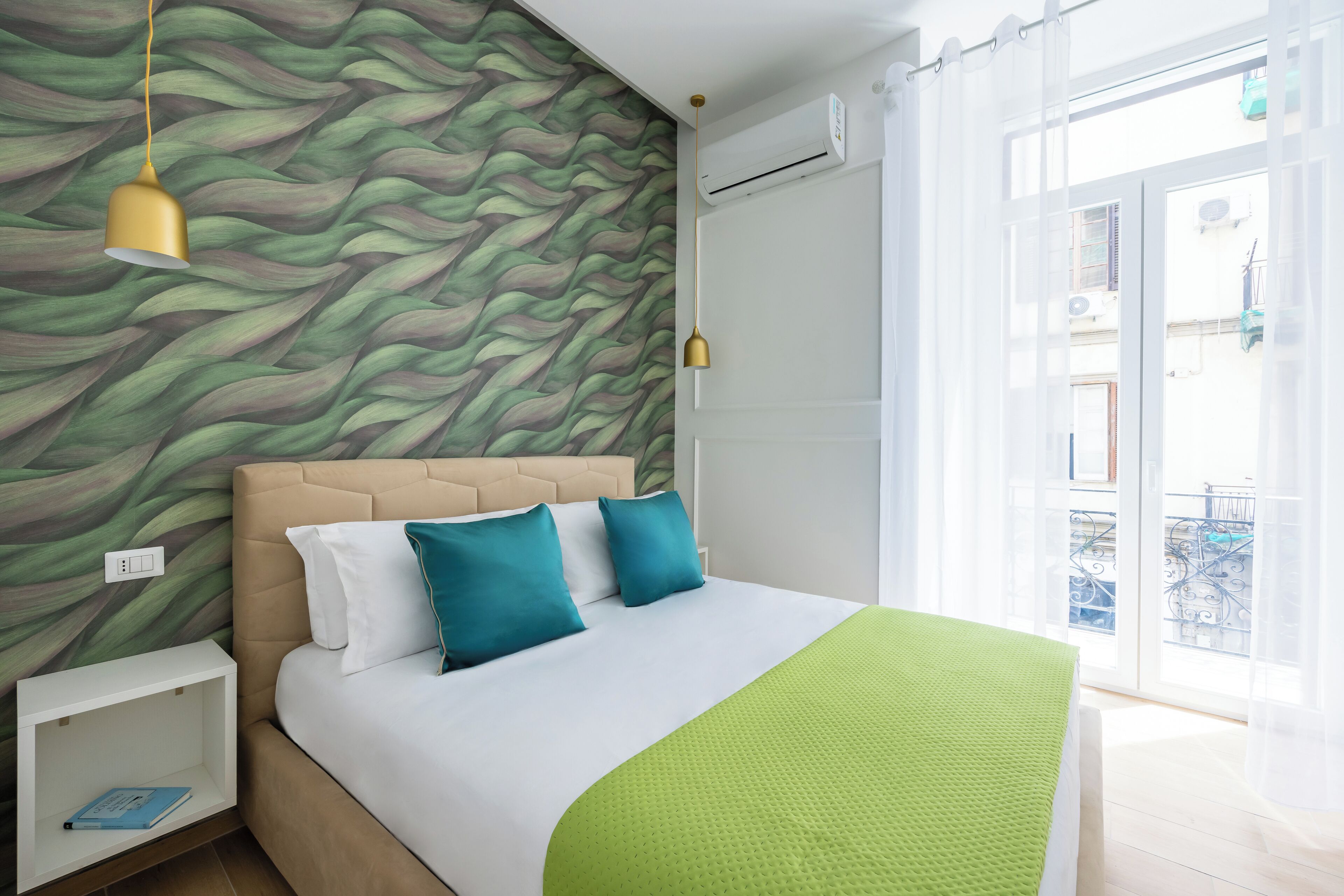 Un lit bien fait, recouvert d’une couverture verte, avec une tête de lit et deux oreillers. On y trouve une table de chevet sur laquelle sont posés un livre et une lampe. La pièce est décorée d’un mur à motifs et possède une fenêtre avec des rideaux.