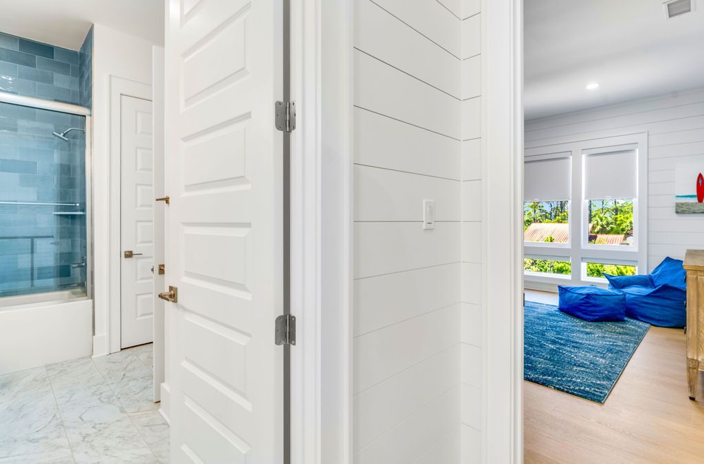 A hallway with a glass shower, white doors, and a view of a living area with a blue rug and wooden furniture.