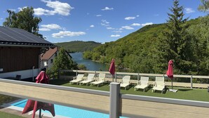 Un área junto a la piscina con sillones de descanso y sombrillas, con vistas a un lago y colinas boscosas.