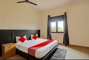Un dormitorio con cama, mesita de noche, ventilador de techo, ventana con cortinas y vista a follaje exterior.