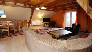Un salon chaleureux de style industriel, avec un plafond haut, des poutres en bois, un canapé, une table à manger et un téléviseur.