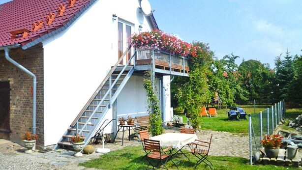 Une maison blanche avec un toit rouge, un balcon orné de fleurs et un escalier menant à l’entrée.