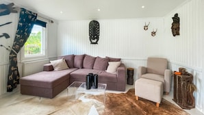 Un salon comprenant un canapé d’angle, un petit fauteuil, une table basse en verre, un tapis en fourrure et des éléments décoratifs muraux, notamment des crânes d’animaux.