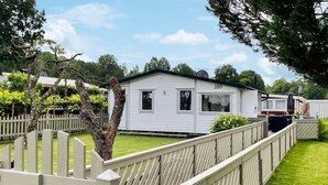Une maison mobile blanche, avec le numéro « 100 » peint sur le côté, entourée d’une clôture en bois et d’une végétation luxuriante.