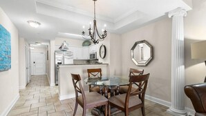 A dining area with a glass table surrounded by chairs, a chandelier, and a kitchen in the background. - Oceanfront Paradise Perfection 2BR Condo/ Balcony (North Myrtle Beach)