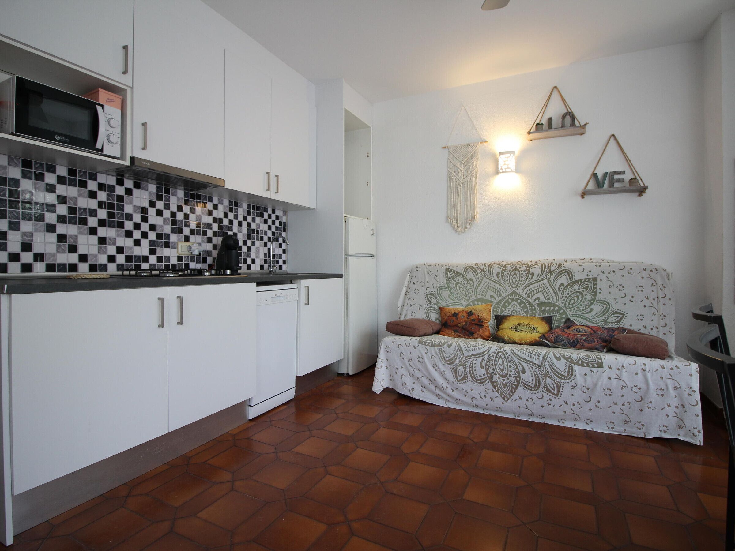 A compact kitchen with white cabinets, a microwave, and a dishwasher. There is a patterned sofa with cushions, a wall-mounted light, and a decorative macrame wall hanging.