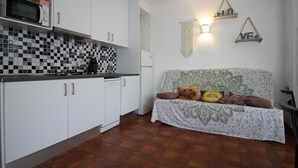 Eine kompakte Küche mit weißen Schränken, einer Mikrowelle und einem Geschirrspüler. Es gibt ein gemustertes Sofa mit Kissen, eine Wandleuchte und ein dekoratives Makramee-Wandbild.
