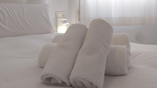 Een keurig opgemaakt bed met opgerolde witte handdoeken, een bedlamp en dunne gordijnen.