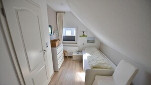 A small, well-lit room with a bed, a chair, a dresser, and a window.