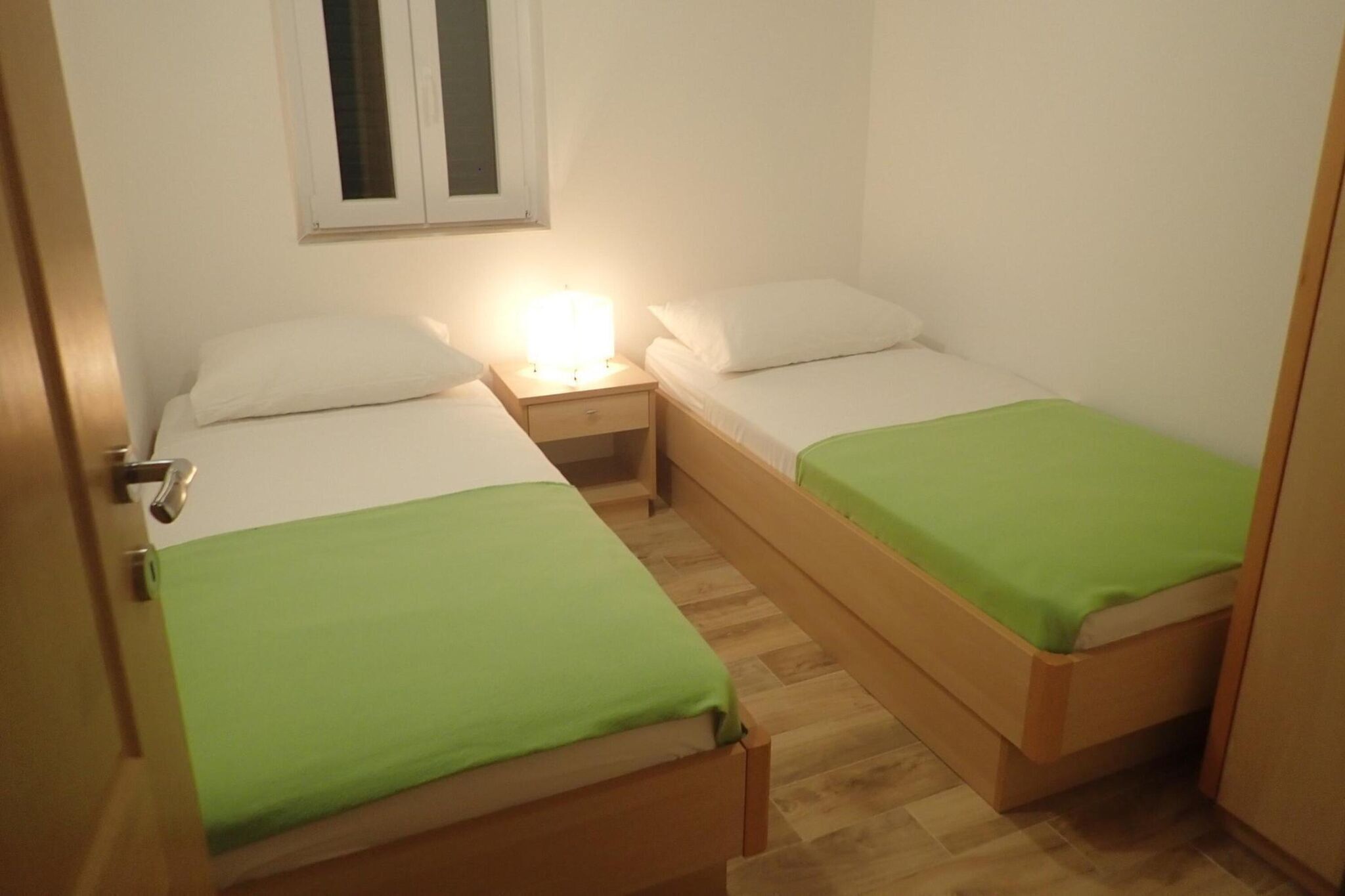 A small, compact room with two single beds, each with a green bedspread. There is a nightstand with a lamp between the beds, and a window above the left bed.