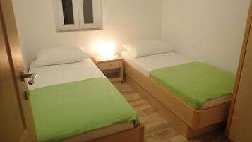 A small, compact room with two single beds, each with a green bedspread. There is a nightstand with a lamp between the beds, and a window above the left bed.