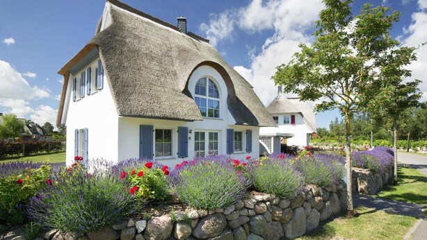 Et hus med stråtækt tag, hvide vægge og en stenvæg med lavendel og røde blomster.