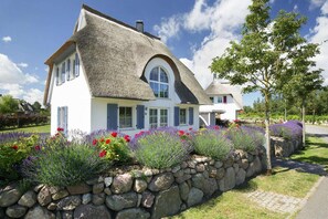 Ein Haus mit Strohdach, weißen Wänden und einer Steinmauer mit Lavendel und roten Blumen.