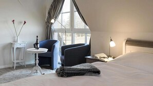 Et hotelværelse med en seng, to stole, et lille bord med en flaske, en vase med blomster, en lampe og et vindue med gardiner.