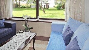 A living room with a blue sofa, a coffee table with a vase of white flowers, and a view of a grassy garden through a large window. - Holiday Home Sulomino with Private Beach (Sulomino)