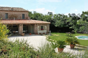 A rustic stone house with a tiled roof, a patio with outdoor furniture, and a swimming pool.