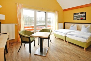 Habitación de hotel con una cama, una mesa, dos sillas, una ventana con cortinas, un radiador y un cuadro en la pared.