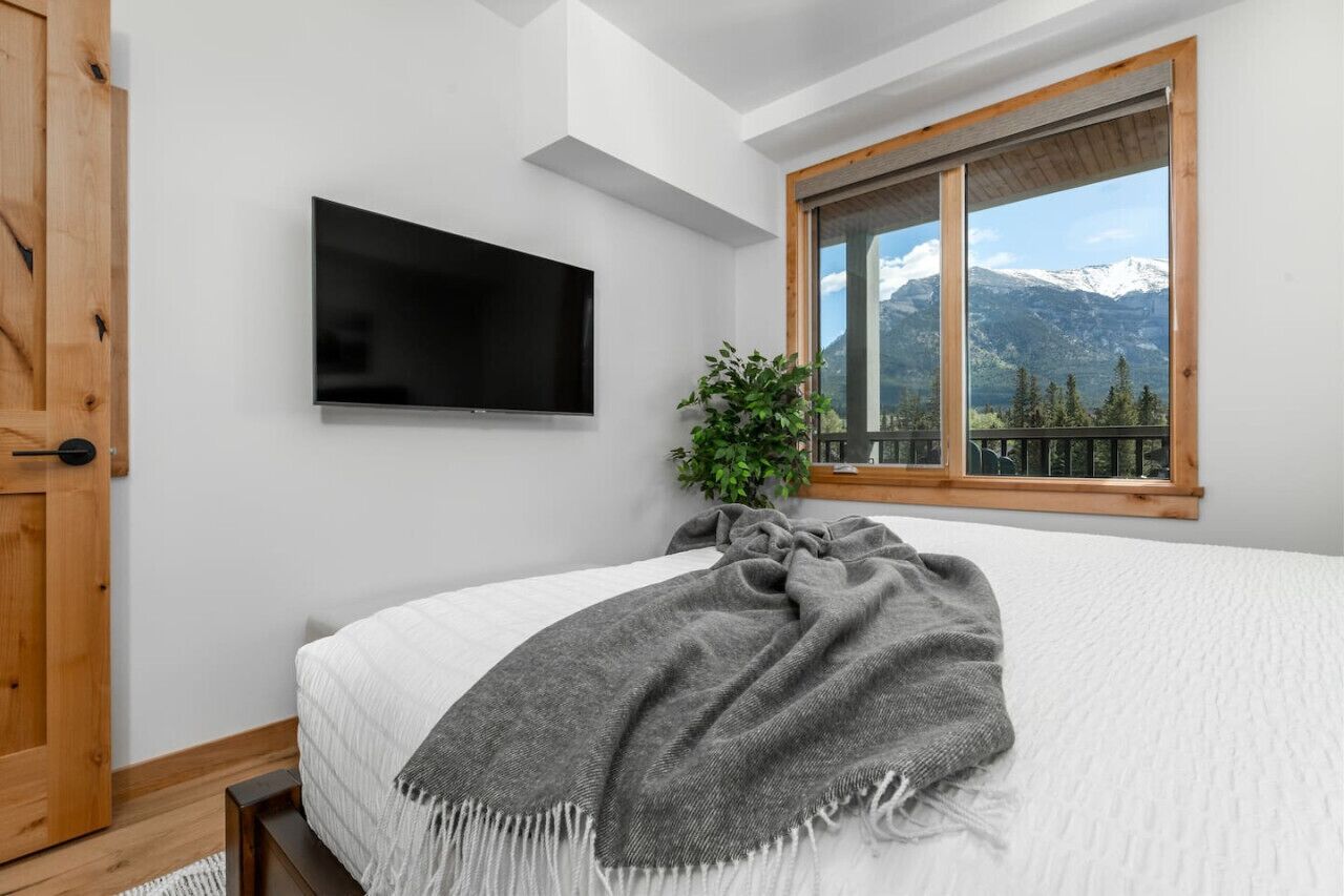 A bedroom with a bed, a television mounted on the wall, a window with a mountain view, and a wooden door.