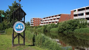 A sign with a sailboat and text, a body of water, and a building with multiple balconies.