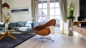 A modern living room with a sofa, a brown chair, a glass coffee table, a TV, and a view of a house through the window. - Reetdachhaus am Bodden mit Sauna (Fuhlendorf)