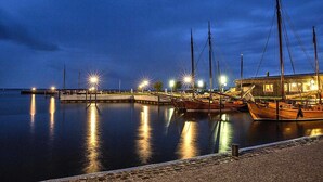 A marina at dusk with several boats docked, a wooden building with lights, and a calm water surface reflecting the lights. - Reetdachhaus am Bodden mit Sauna (Fuhlendorf)